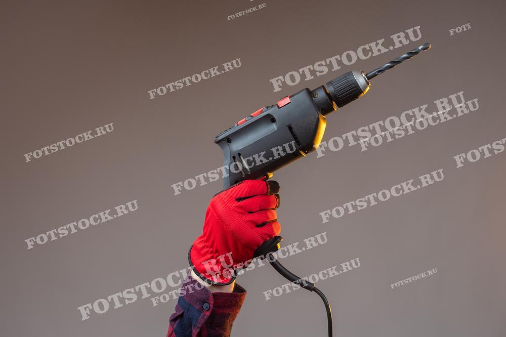 На фотографии изображена рука в красной рабочей перчатке, держащая электрическую дрель с установленным сверлом. Такой снимок может быть использован в рекламных каталогах инструментов, обучающих материалах по работе с электроинструментами, статьях о ремонте и строительстве, а также в магазинах DIY (сделай сам).
