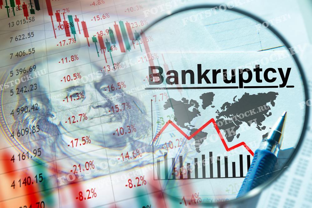 На изображении лупа показывает слово "Bankruptcy" на фоне падающих графиков и купюры с изображением Бенджамина Франклина. Эта композиция символизирует финансовый крах и экономический спад. Графики и денежные знаки подчеркивают серьезность ситуации и масштаб кризиса.