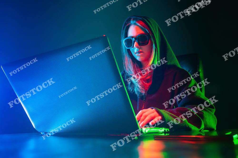 На фотографии изображена женщина в темной одежде, очках и худи, работающая за ноутбуком. Освещение в синих и зеленых тонах подчеркивает интригующую и высокотехнологичную атмосферу. Образ подходит для иллюстрации тем, связанных с кибербезопасностью, технологиями или программированием.