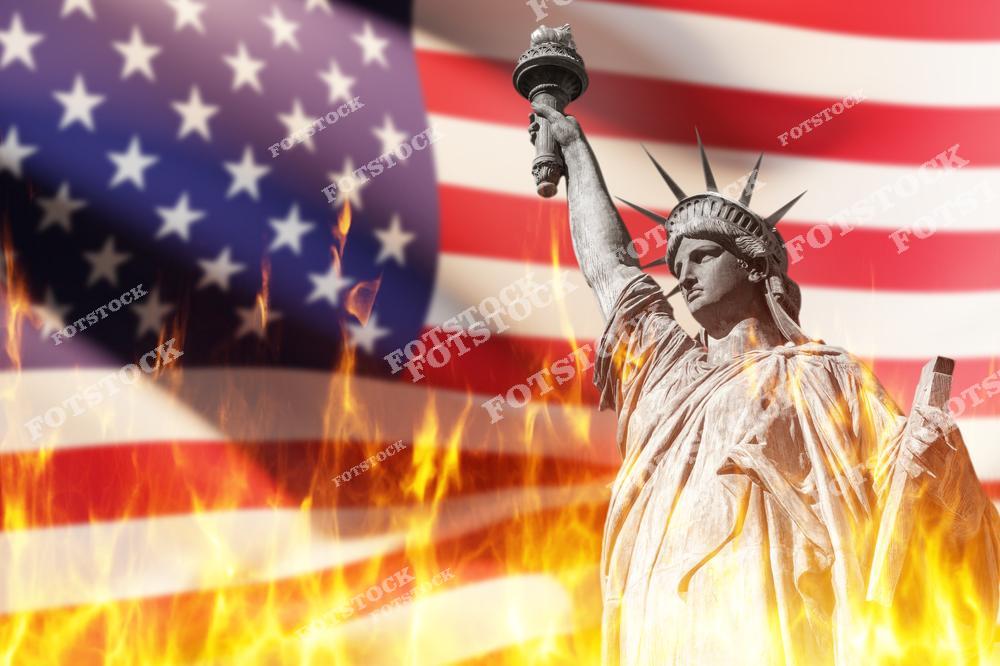 На изображении Статуя Свободы на фоне развевающегося флага США, окруженная пламенем. Композиция передает драматизм, напряжение и вызовы, с которыми сталкивается Америка.