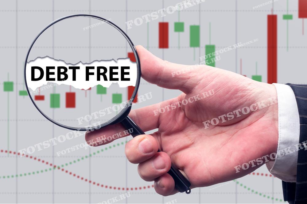 Фотография показывает руку, держащую лупу, через которую видно надпись "Debt Free" на фоне финансовых графиков. Это изображение символизирует достижение финансовой свободы и анализ текущего финансового состояния.