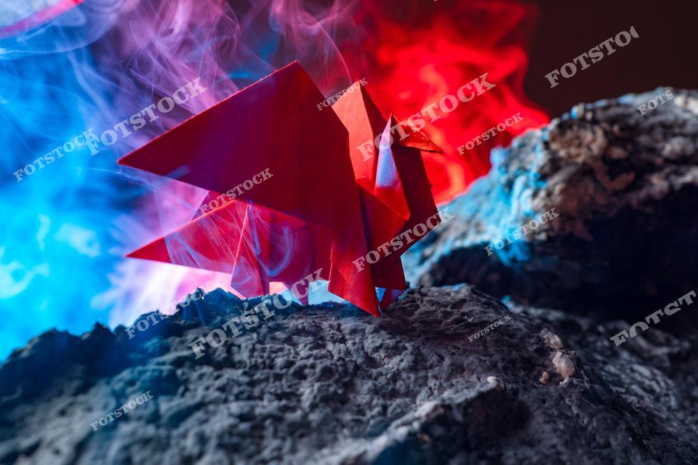 Фотография изображает красную оригами-фигуру дракона, стоящего на скале среди густого дыма с подсветкой синего и красного цветов. Композиция напоминает кадр из фантастического фильма или игры. Атмосфера загадочна и наполнена символикой мифов и волшебства.