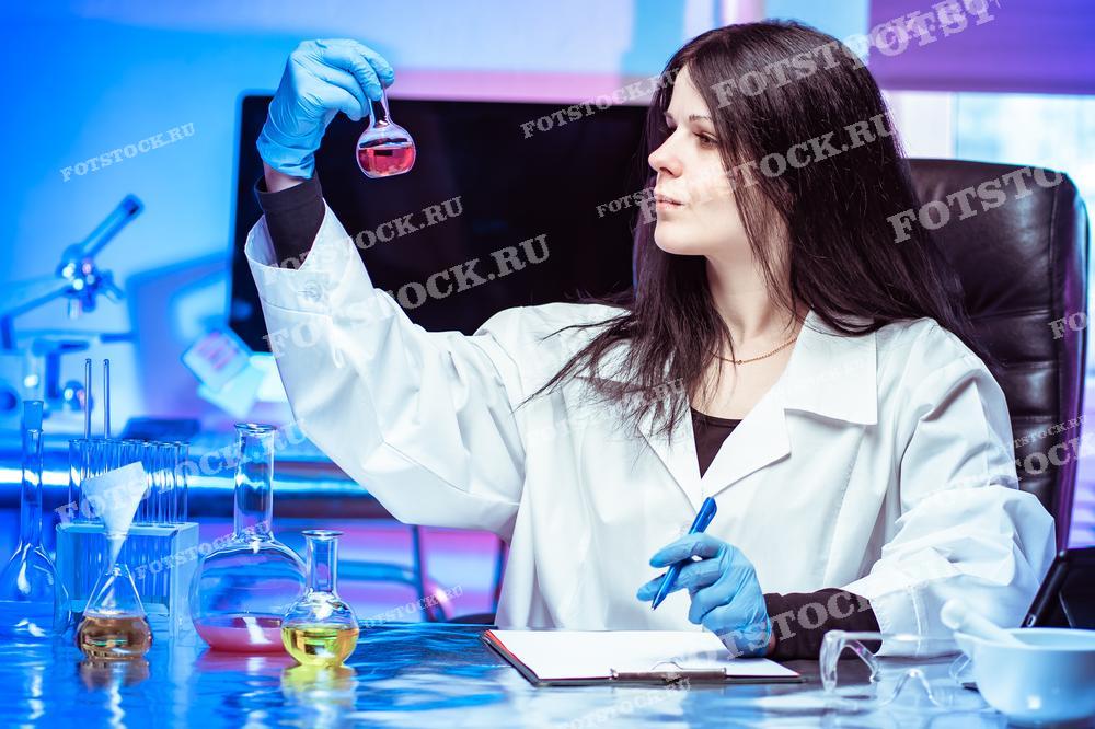 На изображении женщина в белом лабораторном халате и перчатках внимательно изучает прозрачный флакон с красной жидкостью на фоне синей подсветки лаборатории. Это изображение может быть использовано для научных публикаций, промо материалов для лабораторий, а также в учебных целях.