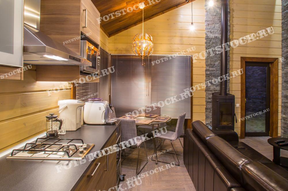 На фотографии изображена современная кухня-гостиная в загородном доме. Интерьер выполнен в теплых тонах с использованием натуральных материалов, таких как дерево и камень. Кухонное пространство оборудовано современной техникой, а рядом находится обеденная зона с уютным столом и креслами. В комнате также установлен камин, создающий атмосферу тепла и уюта.