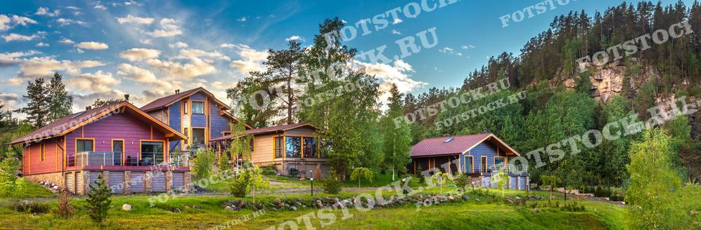 На фотографии изображены загородные дома, расположенные в живописной местности, окруженные лесом и горами. Летний пейзаж с зелеными деревьями и ярким небом создает ощущение спокойствия и гармонии с природой. Эти дома предлагают идеальные условия для комфортного и уединенного отдыха на свежем воздухе вдали от городской суеты.