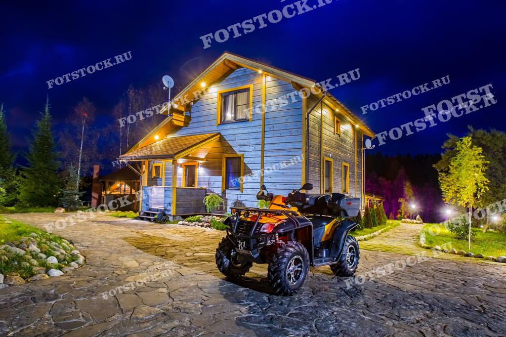На фотографии изображен загородный дом в ночное время, с включенной внешней подсветкой, что создает атмосферу уюта и комфорта. Перед домом на каменной подъездной дорожке стоит квадроцикл, готовый к приключениям на природе. Вокруг дома можно увидеть деревья и зелень, подчеркивающие гармонию с окружающей средой.