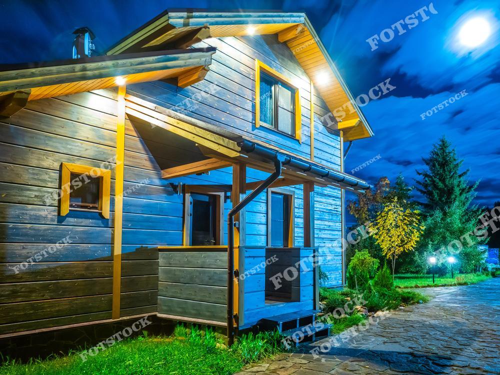 Фотография демонстрирует деревянный дом с террасой в ночное время. Освещение дома и лунный свет подчёркивают его архитектуру на фоне синего облачного неба. Уютный загородный пейзаж и спокойствие природы создают гармонию.