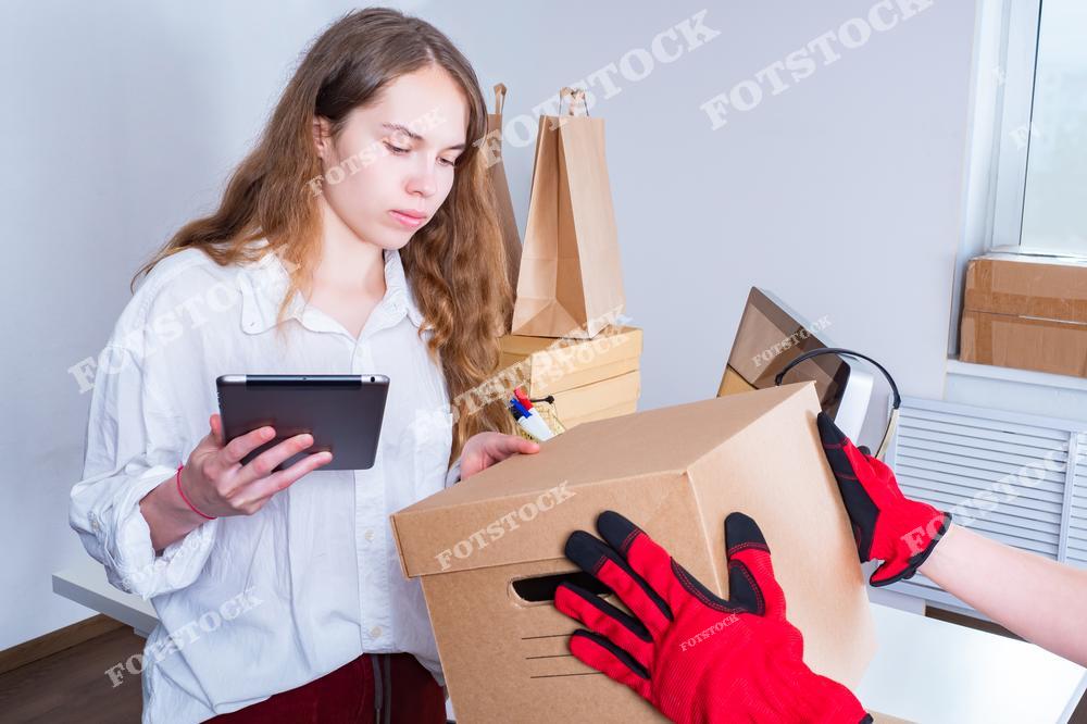 На изображении девушка с планшетом в руках принимает доставленную картонную коробку от курьера в красных защитных перчатках. На заднем плане видны другие упакованные коробки и бумажные пакеты. Сцена иллюстрирует процессы электронной коммерции и подтверждения получения товара.