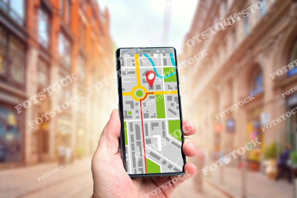 На переднем плане изображена рука, держащая смартфон с картой и указателем местоположения. На фоне видны городские улицы и здания, создавая атмосферу динамичного городского окружения. Фотография подчёркивает использование современных технологий навигации для ориентации и передвижения по городу.