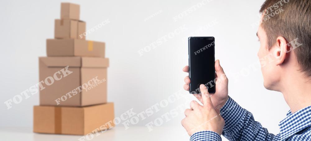 На изображении мужчина в клетчатой рубашке держит в руках смартфон, направляя его в сторону пирамиды из картонных коробок. Сцена символизирует современные процессы в сфере логистики и доставки, где цифровые технологии позволяют отслеживать и управлять грузами в реальном времени.