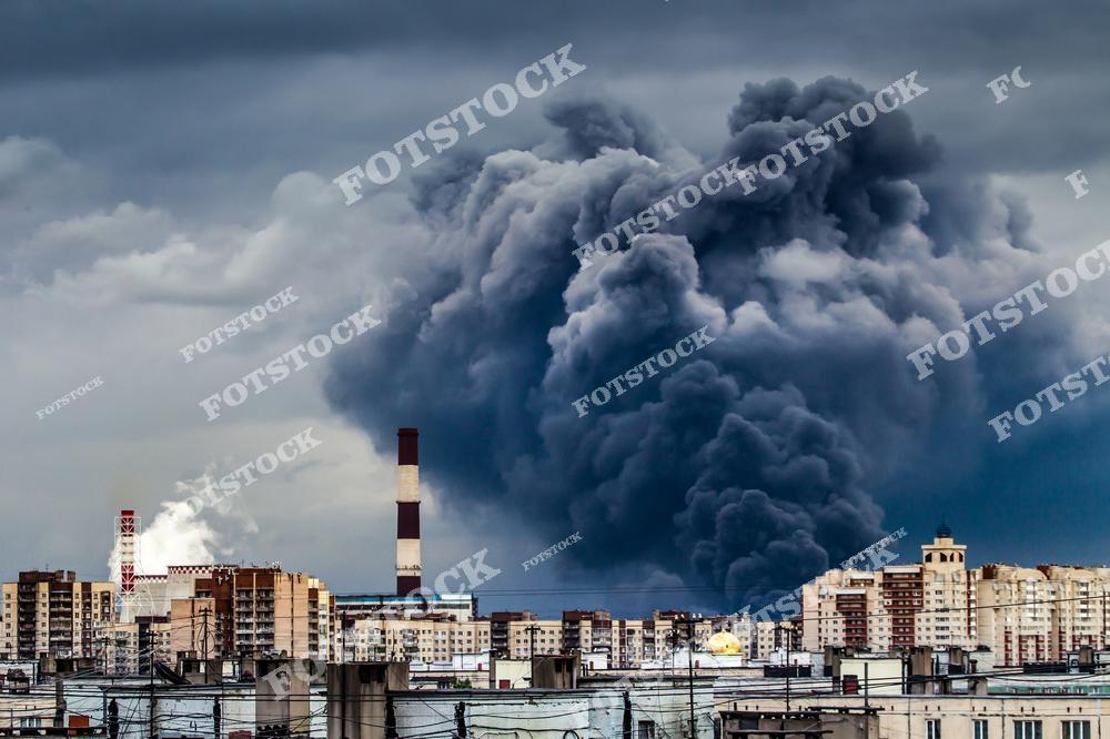 На фотографии изображен густой черный дым от пожара, поднимающийся над жилыми домами. На фоне видны промышленные трубы, что подчеркивает связь техногенной деятельности с экологическими проблемами и загрязнением воздуха.