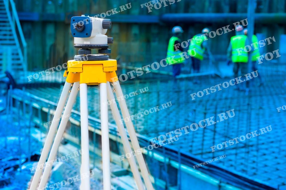 На фотографии изображен нивелир, установленный на треноге на строительной площадке. На заднем плане видны рабочие в ярких жилетах, выполняющие работы по заливке фундамента. Изображение подчеркивает важность точных измерений в строительстве.