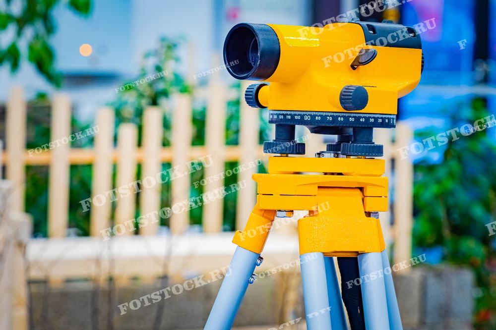 Фотография демонстрирует желтый геодезический нивелир, установленный на штативе, готовый к работе. На заднем плане виден размытый забор и растительность, что подчеркивает использование нивелира в условиях открытой местности или на строительной площадке.