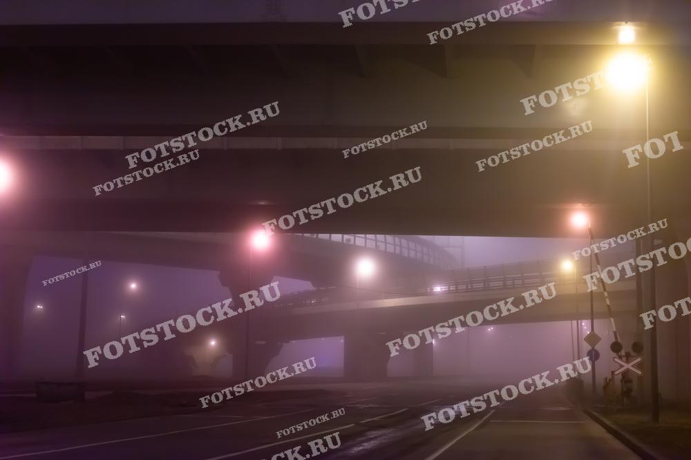 Ночная фотография мостовой развязки, окутанной густым туманом, с приглушенным освещением уличных фонарей. Изображение создает мистическую атмосферу и подчеркивает особенности городской инфраструктуры в условиях плохой видимости.