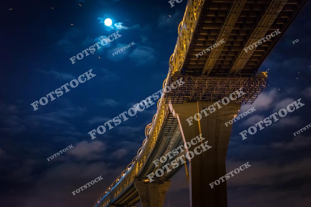 На фотографии запечатлен мост с массивными опорами, подсвеченный искусственным светом, на фоне глубокого ночного неба. Яркая луна сияет сквозь облака, придавая картине загадочную и драматичную атмосферу. Внизу видны детали металлической конструкции, подчеркивающие инженерное мастерство.