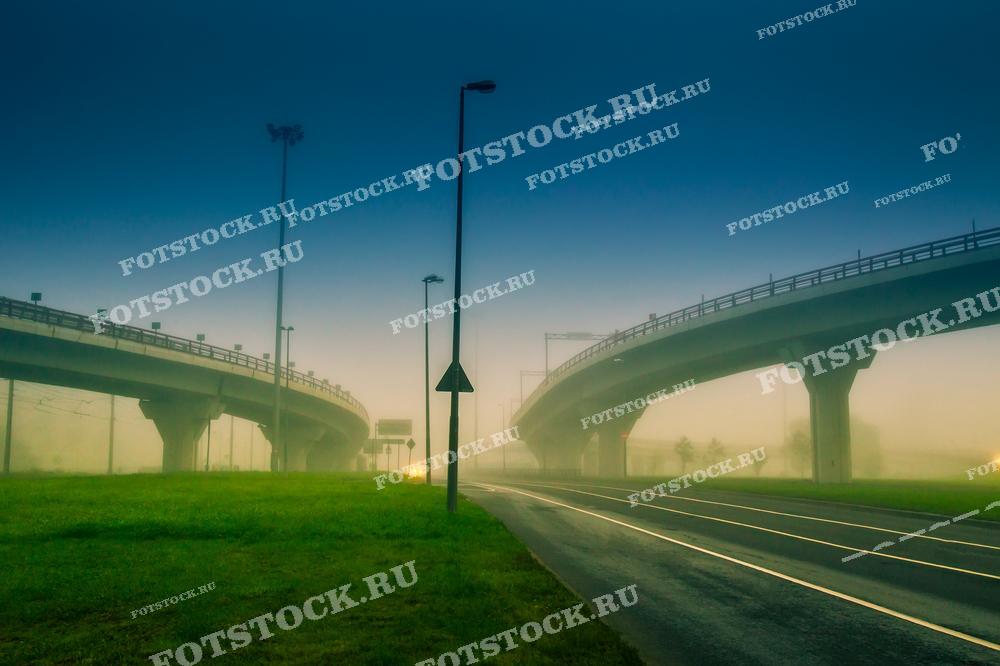 Фотография мостовой развязки, погруженной в туман на рассвете. Снимок создает ощущение тишины и загадочности, подчеркивая контраст между зеленой травой и бетонными конструкциями в условиях слабой видимости.