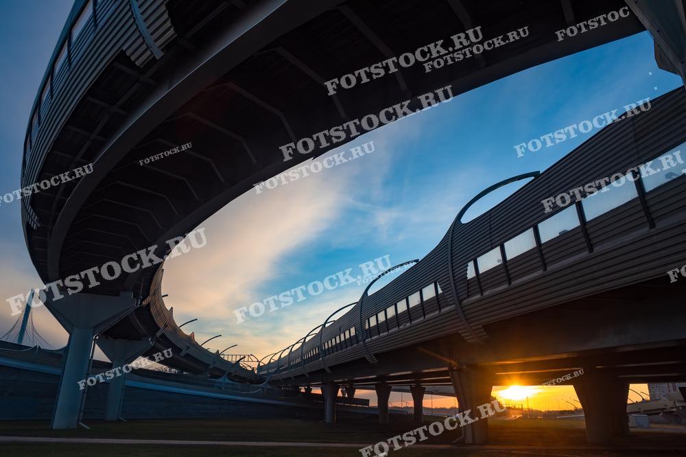 Фотография мостовой развязки, освещенной закатным светом. Снимок подчеркивает величие и изящество городской архитектуры на фоне яркого неба и уходящего солнца.