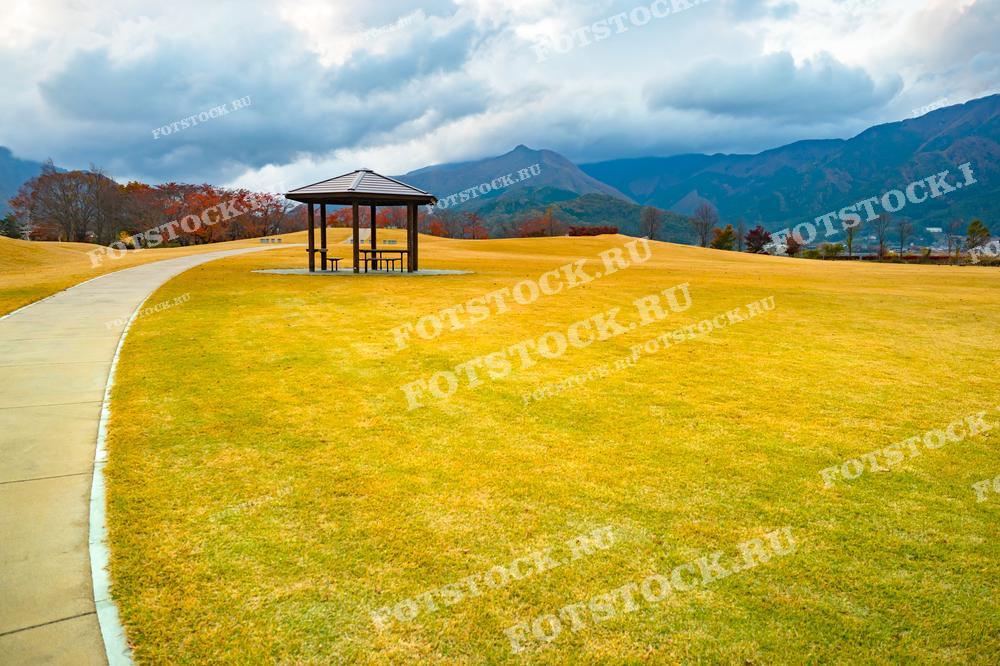 На фотографии изображена тропинка в японском парке Ягидзаки, которая ведёт к озеру Кавагутико. В центре парка расположена беседка, окружённая ярко-жёлтой травой, на фоне осенних деревьев и гор деревни Фудзикавагутико. Облачное небо добавляет спокойную и умиротворённую атмосферу осеннего дня.