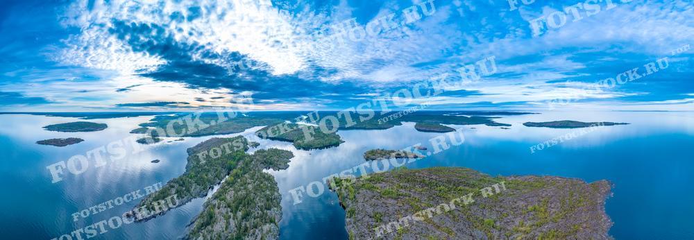На фотографии представлена панорама Ладожских шхер, снятая с высоты. Вид открывается на многочисленные острова и полуострова, омываемые спокойными водами Ладожского озера. Плотные облака на небе и их отражение в воде создают впечатление безграничного простора. Ладожские шхеры — это уникальный природный комплекс, привлекающий множество туристов своими живописными видами и возможностью уединенного отдыха. Эта панорама подчеркивает всю красоту и величие северной природы Карелии.