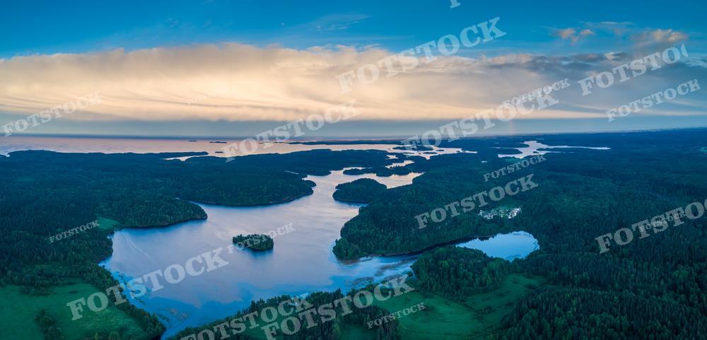 Эта фотография показывает панорамный вид на водоёмы и леса Карелии в мягком свете восхода или заката. Сложная система озёр, проток и островов сочетается с тёмно-зелёной растительностью, а небо покрыто тяжёлыми облаками, отражающими свет солнца. Изображение может быть использовано для научных исследований, экологических презентаций, в качестве фона для медитативных практик или для украшения статей о путешествиях и природе.
