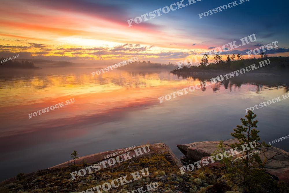 На фотографии запечатлён рассвет над Ладожским озером в Карелии. Небо и вода переливаются золотистыми и розовыми оттенками, отражая первые лучи солнца. Спокойная поверхность озера и скалистый берег создают атмосферу умиротворения и красоты. Этот вид подчёркивает природное великолепие Карелии, делая её популярным местом для любителей природы.