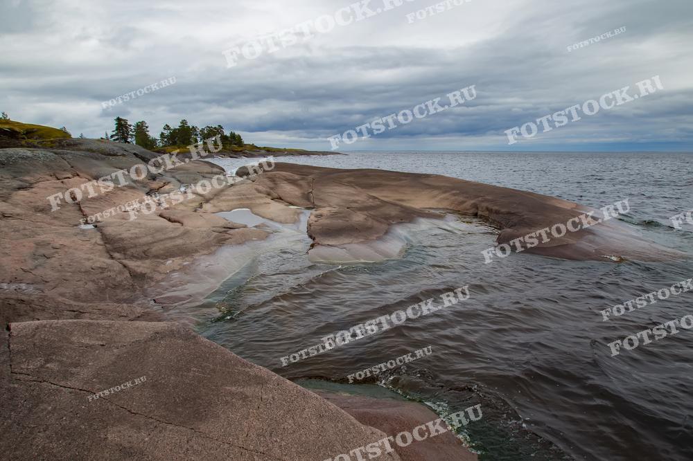 На фотографии изображены скалистые берега Ладожского озера, одного из крупнейших пресных озер Европы. Каменные формы береговой линии создают необычный и живописный пейзаж. Местность характеризуется гладкими и выветренными скалами, плавно опускающимися в воду. Ладожские шхеры — это популярное место среди туристов, особенно для тех, кто любит дикий отдых на природе, походы и водные прогулки.