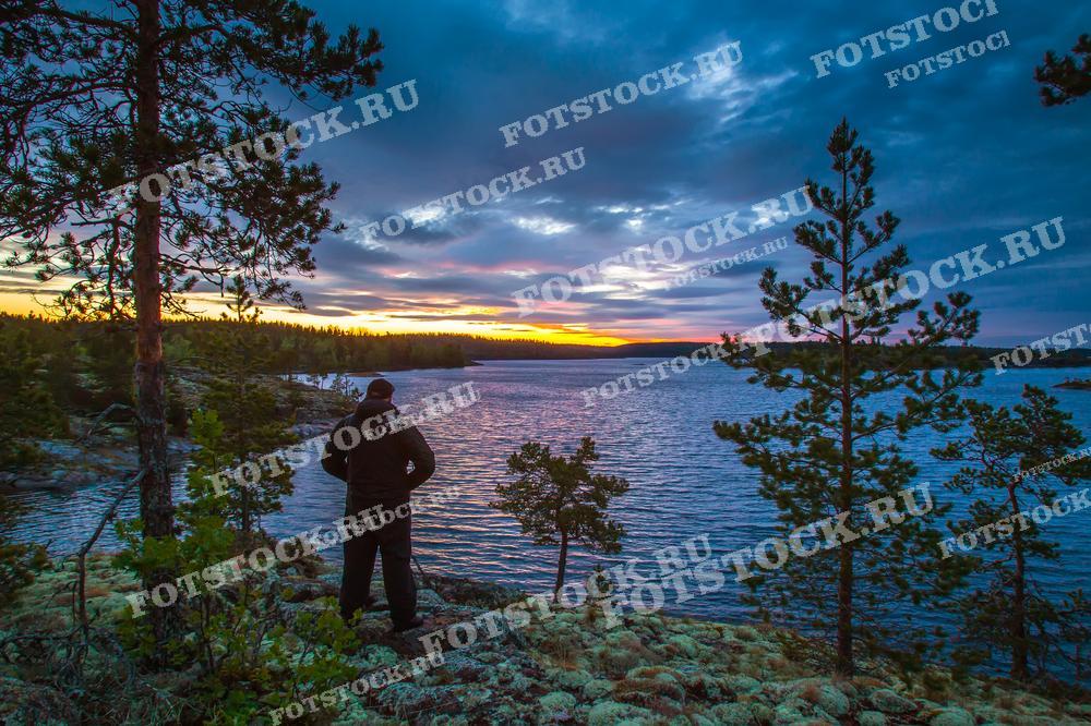 На фотографии изображен мужчина, стоящий на берегу озера в Карелии. Вечернее небо, окрашенное закатными красками, отражается в воде, создавая неповторимую атмосферу. На переднем плане видны сосны и каменистый берег, типичный для карельских шхер. Этот вид характерен для Ладожского озера, крупнейшего озера Европы, которое славится своей живописной природой и является популярным местом для отдыха и туризма.