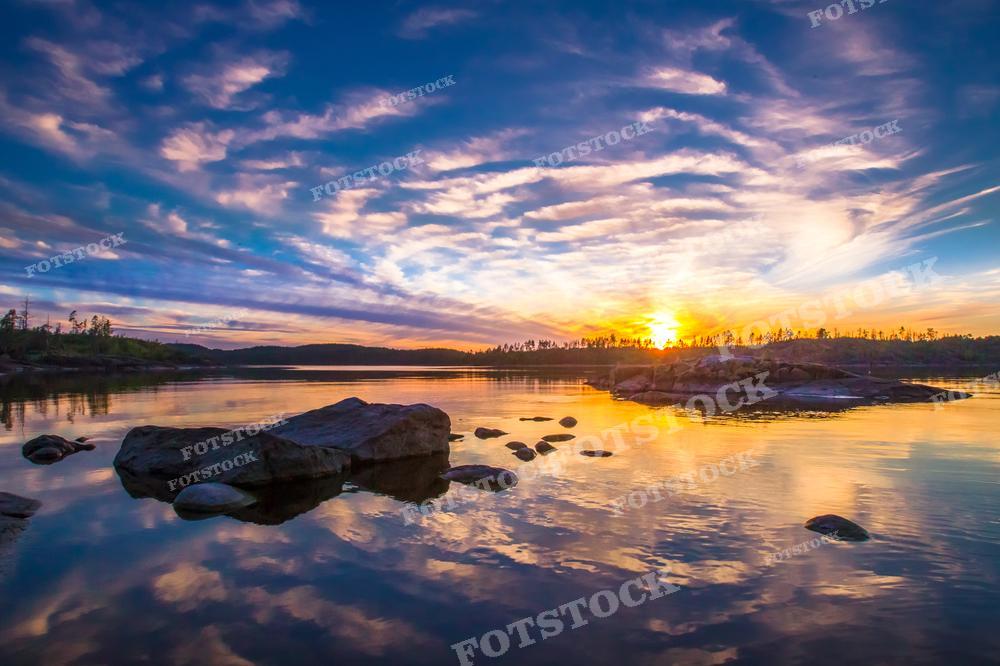 На фотографии изображен заход солнца над озером в Карелии. Вода зеркально отражает разноцветные облака, которые раскинулись по небу. Видны камни в воде на переднем плане и лесистый берег на заднем. Солнце, приближающееся к горизонту, создаёт тёплые тона на небе и на поверхности воды, придавая сцене спокойствие и торжественность. Такая фотография может быть использована в туристических проспектах, для оформления статей о природе, как элемент дизайна интерьеров или в качестве обоев для рабочего стола.