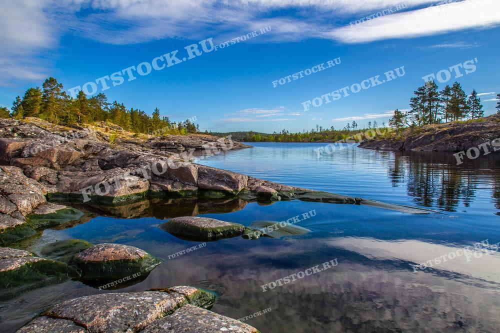 На фотографии изображен берег Ладожского озера, где камни и спокойная вода создают впечатляющий пейзаж. Голубое небо с легкими облаками отражается в озере, добавляя снимку глубины и яркости. На заднем плане видны густые зеленые леса, которые характерны для карельской природы. Это место идеально подходит для туристов, ищущих уединение и возможность насладиться красотой природы.