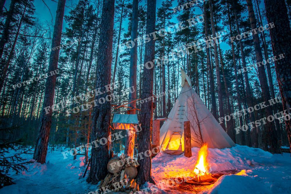 На фотографии изображён зимний лес в Карелии, где среди высоких сосен стоит белая палатка в стиле вигвама. Перед палаткой горит костёр, создавая теплое и уютное освещение. Снег вокруг палатки и на деревьях подчёркивает зимнюю атмосферу, а сумерки придают пейзажу таинственности и очарования. Эта сцена демонстрирует идеальное место для зимнего отдыха на природе, в окружении спокойствия и красоты карельского леса.