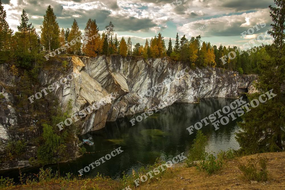 На фотографии изображён знаменитый мраморный карьер в горном парке Рускеала в Карелии. Бело-серые мраморные скалы подчёркивают природное величие этого места, а осенние краски делают пейзаж ещё более живописным. Горный парк Рускеала — это популярное туристическое направление, известное своими мраморными каньонами, озёрами и шахтами. Это место привлекает путешественников в любое время года, но осенью оно выглядит особенно красиво благодаря ярким цветам природы.