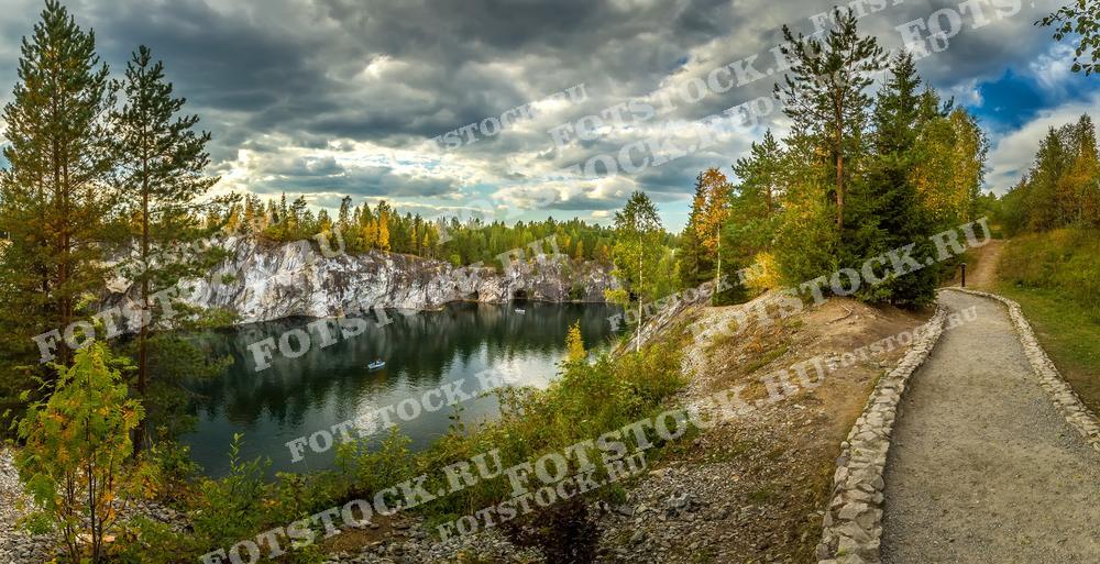 На фотографии запечатлен красочный осенний пейзаж Карелии. Изображен уединённый карьер с кристально чистой водой, окружённый разноцветной растительностью и высокими хвойными деревьями. Прокладываемая каменная тропа ведёт к этому живописному месту, предлагая идеальный маршрут для прогулок на свежем воздухе. Это место может быть популярным среди туристов, ищущих уединение и контакт с природой, а фотография подходит для иллюстрации статей о природных достопримечательностях или в рекламных брошюрах туристических агентств.