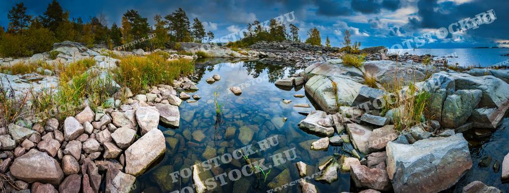 На фотографии изображен панорамный вид на берег Ладожского озера в Карелии. Скалистый и каменистый ландшафт с разнообразной растительностью и чистыми водоемами создает живописную природную сцену. Небо частично покрыто облаками, которые отражаются в воде, придавая изображению глубину и масштаб. Этот кадр подчеркивает уникальную красоту северной природы и может использоваться в туристических брошюрах, экологических публикациях или как элемент декора.