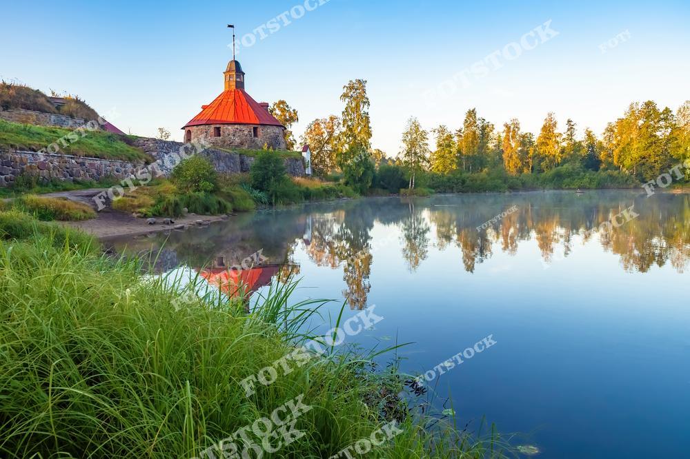 На фотографии изображена крепость Корела, расположенная в городе Приозерск, Республика Карелия. Старинные каменные стены и башня с красной крышей отражаются в спокойной воде реки. Утренний свет освещает зелень вокруг, подчёркивая гармонию архитектуры и природы. Крепость Корела — историческая достопримечательность, которая сыграла важную роль в защите северо-западных рубежей России.