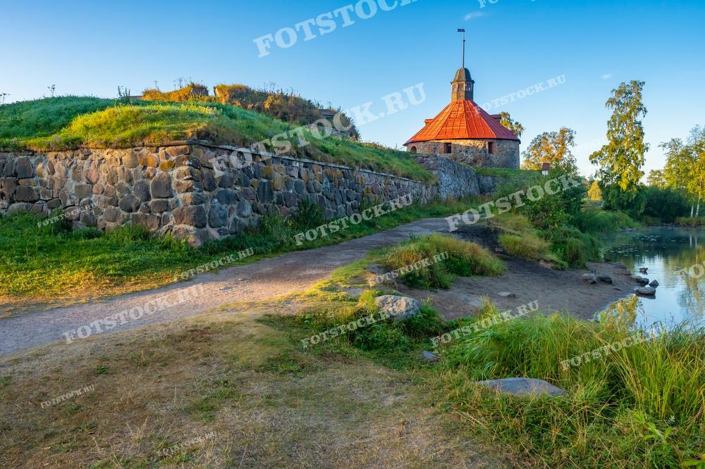 На фотографии изображен вид на Крепость Корела в Приозерске, Карелия. Крепостная стена из натурального камня и земляной насыпи ведет к башне с красной крышей, которая является центральным элементом композиции. Тихая речка у подножия крепости добавляет покой и умиротворение к сцене, делая это место идеальным для исторических экскурсий и пейзажной фотографии.