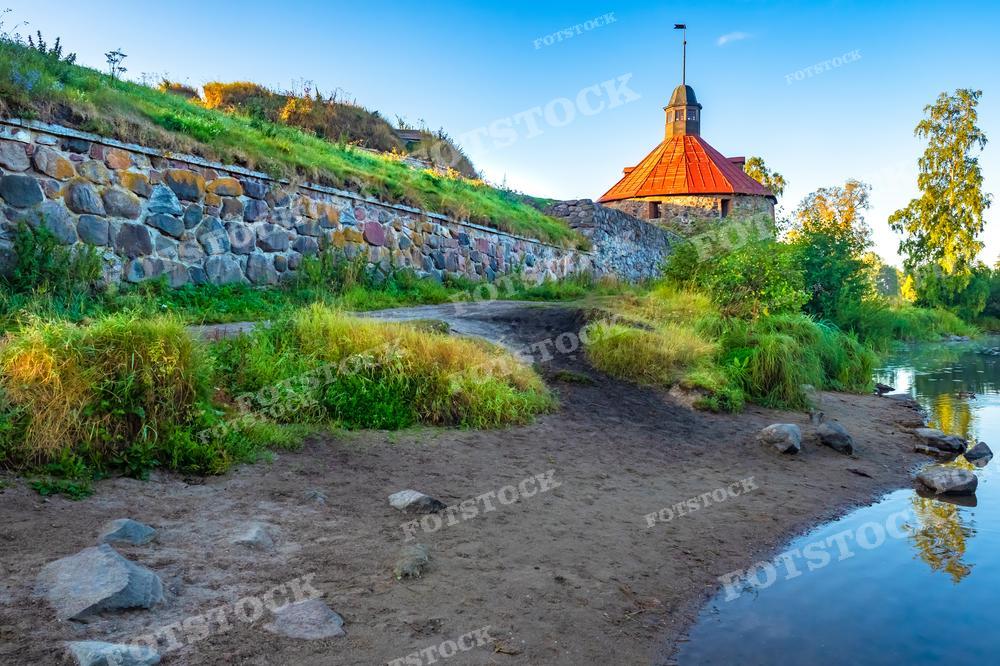 На фотографии изображен старинный каменный бастион или часть крепости, расположенный на берегу в Карелии. Зеленые насаждения украшают стены, а рядом с бастионом протекает небольшая река или ручей. Это теплое и мирное утро, и лучи солнца освещают каменную стену и покрывают золотом прибрежные кусты. Такое изображение подходит для исторических и культурных публикаций, может быть использовано в туристических брошюрах или как фото для статей о местной архитектуре.