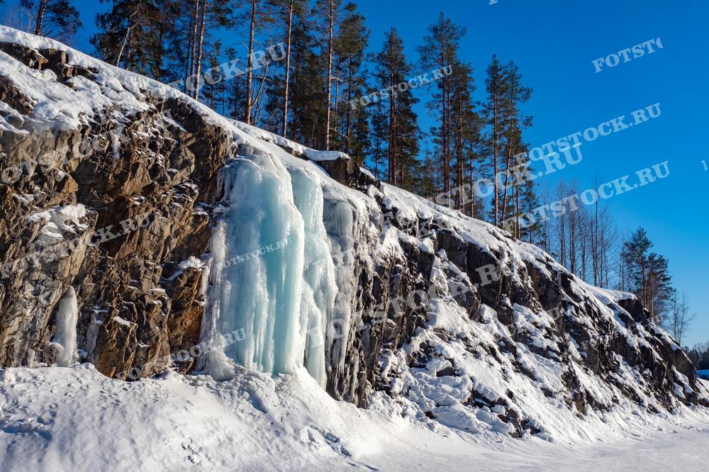 На фотографии изображены скалы, покрытые льдом и снегом, в зимней Карелии. Ледяные сосульки свисают с каменистых обнажений, создавая впечатляющий контраст с синим небом и сосновым лесом на заднем плане. Яркое зимнее солнце освещает этот живописный пейзаж.