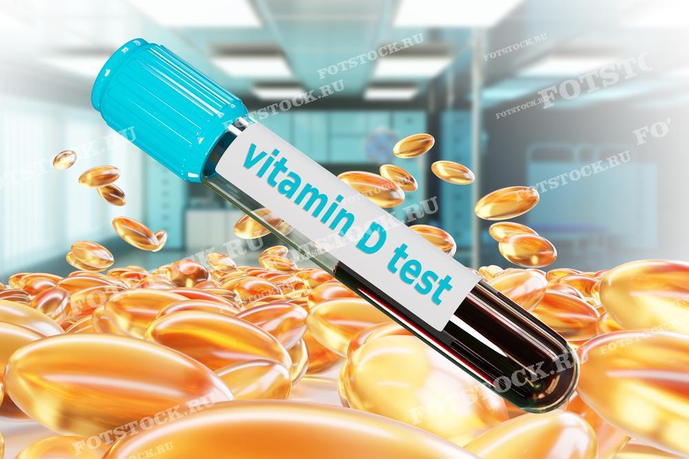 На изображении представлена пробирка с надписью "vitamin D test", окруженная капсулами с витаминами, на фоне лабораторного помещения. Фотография символизирует медицинское исследование и контроль уровня витамина D в крови.