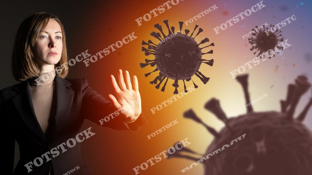 На изображении женщина в деловом костюме символически протягивает руку в защитном жесте, как бы останавливая вирус, визуализированный в форме крупных вирусных частиц с шиповидной структурой на фоне. Это изображение олицетворяет человеческую решимость противостоять пандемии, поиски решений и защиты от инфекции. Используется драматическая игра света и теней, которая усиливает эмоциональный посыл борьбы и решимости.