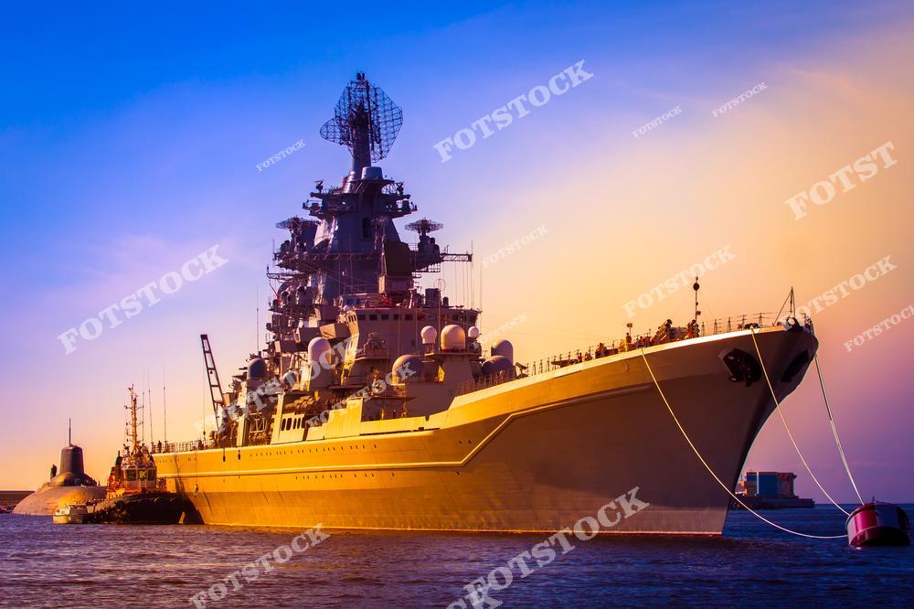 На фотографии изображён крупный военный корабль, пришвартованный рядом с подводной лодкой в лучах заката. Закатное небо и мягкий свет подчёркивают величественность и силу морского флота. Снимок демонстрирует современные военно-морские технологии и торжественную атмосферу.