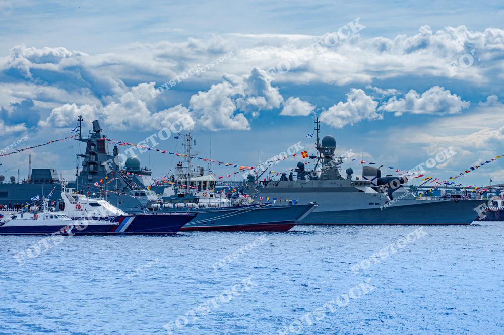 На фото изображены военные корабли и катера, украшенные флагами, на военно-морском параде. На заднем плане видны облачное небо и водная гладь. Фотография символизирует силу флота и торжественные события.