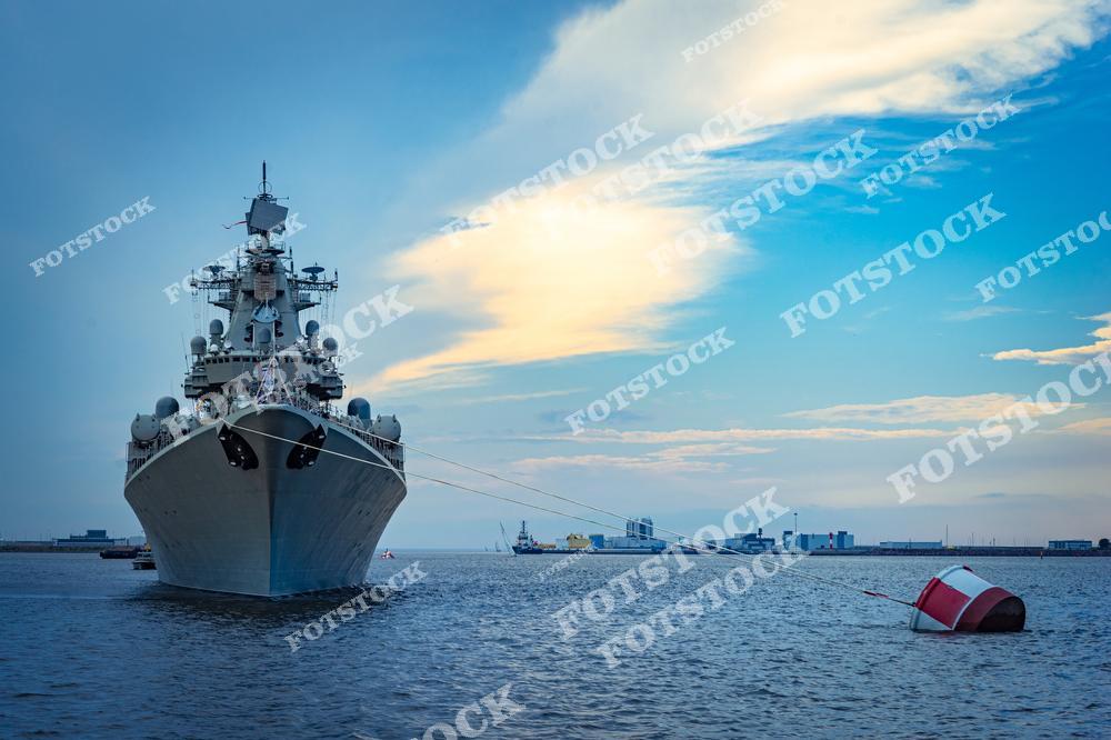 На фото изображен военный корабль, стоящий на якоре в морской гавани, с привязанным к нему буйком. Закатное небо с рассеянными облаками создает спокойное и торжественное настроение. На заднем плане видны элементы портовой инфраструктуры.