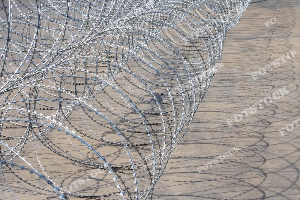На изображении представлена спиральная колючая проволока, выложенная длинной линией на бетонной поверхности. Она символизирует физические заграждения, применяемые для защиты территорий и предотвращения проникновения. Фотография передает строгость и функциональность охранных барьеров.
