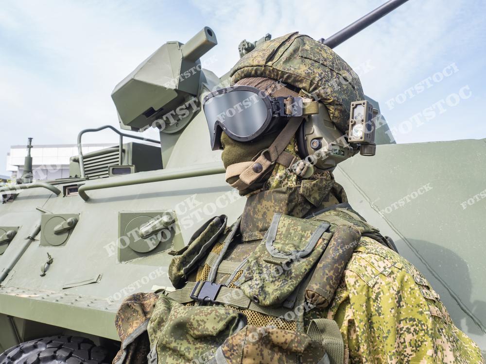 На фотографии изображен военный в камуфляжной экипировке, защитных очках и шлеме, стоящий рядом с бронетранспортером. Оснащение акцентирует внимание на современных технологиях и уровне подготовки. Кадр демонстрирует боевую готовность и тактическое оснащение армии.