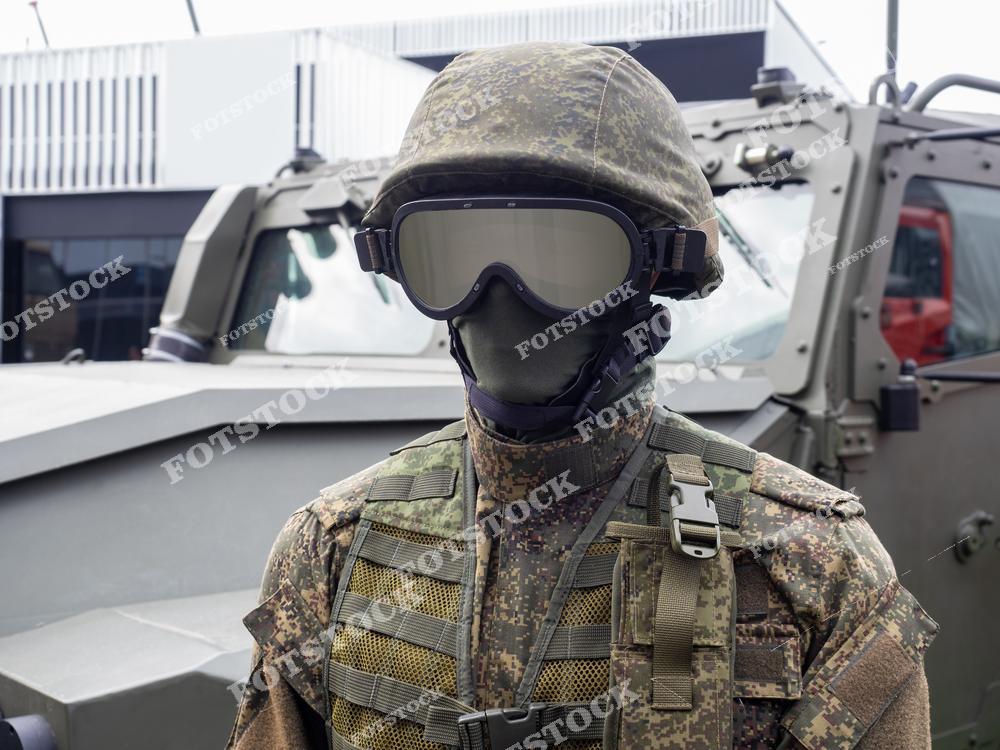 На изображении представлен манекен в военной экипировке с камуфляжным шлемом, защитными очками и тактическим жилетом. На заднем плане виден военный транспорт, подчеркивающий контекст применения оборудования. Фотография иллюстрирует современные технологии и защитные средства для армии.