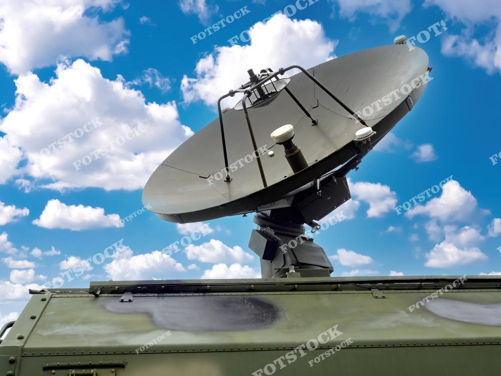 На снимке изображена военная спутниковая антенна, установленная на мобильной платформе. Фон из голубого неба и облаков подчеркивает важность высокотехнологичных решений для связи и радиолокации. Антенна символизирует современные достижения в области военной электроники.