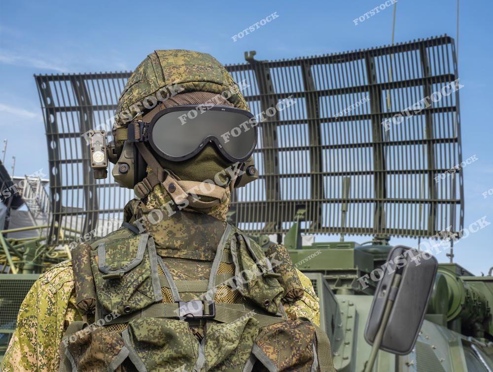 На фотографии изображен военный в камуфляжной форме с защитным шлемом и очками, стоящий на фоне мощной радиолокационной станции. Снаряжение подчеркивает уровень защиты и современные технологии в военном деле. Кадр акцентирует внимание на подготовленности и техническом оснащении вооруженных сил.