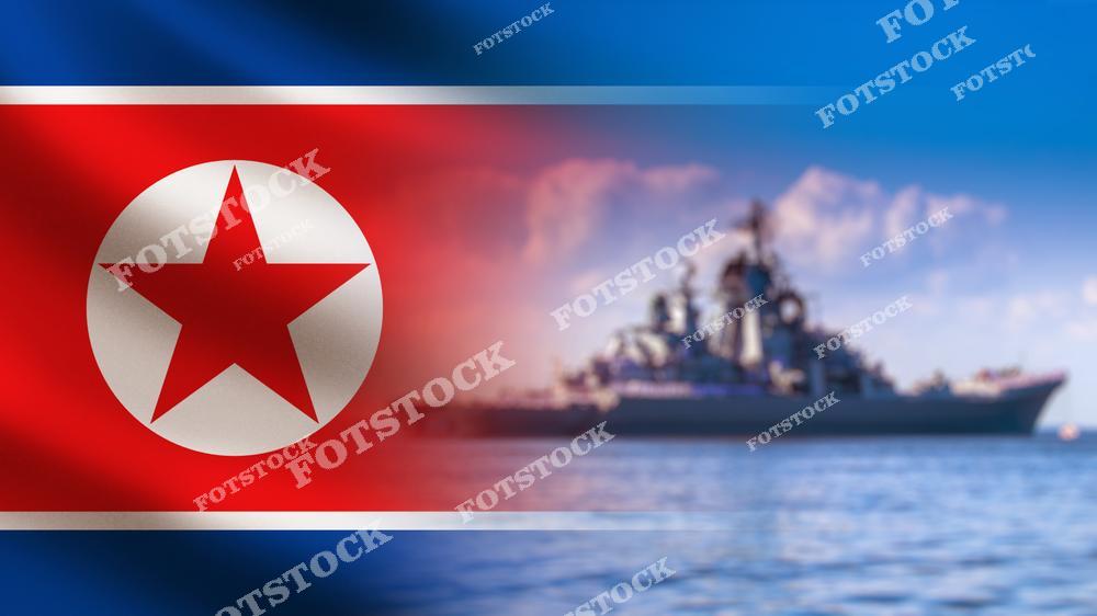 На изображении военный корабль, стоящий на воде, с флагом Северной Кореи на переднем плане. Красный фон с белой звездой подчеркивает национальную символику. Фотография иллюстрирует морскую мощь, стратегическое значение и оборону Северной Кореи.