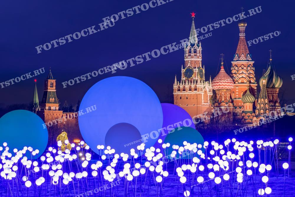 На фотографии изображена Красная площадь с новогодней иллюминацией на переднем плане, Спасская башня и собор Василия Блаженного в Москве. Это фото может быть использовано для иллюстрации праздничного оформления города, в рекламных материалах, посвящённых зимним праздникам, а также для туристических путеводителей.
