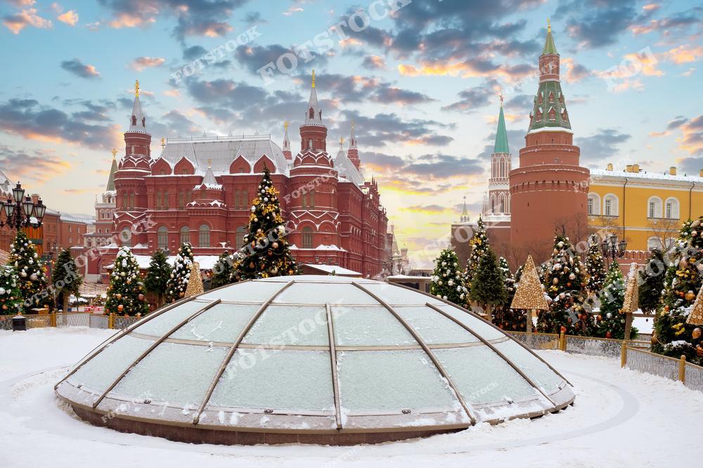 На фотографии изображён зимний пейзаж Красной площади в Москве. На переднем плане — красивые ёлки, украшенные золотыми шарами и гирляндами, создающими атмосферу новогоднего праздника. Вдалеке видны исторические здания, такие как Кремль и здание Государственного исторического музея, которые также дополняют атмосферу зимней Москвы. Снег на земле и холодные голубые оттенки неба придают картине особую магию зимнего времени.